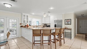 A spacious kitchen with white cabinets, a large island with bar stools, and a view to the outside. - Lakehouse with Sunsets, Private Pool and Jacuzzi, 1Mi to Indian River Lagoon (Sebastian)