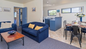 A modern living room with a blue sofa, a wooden coffee table, and a dining area with a table set for four. - Golden Sands Retreat - Matarangi Holiday Home (Matarangi)