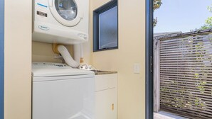 A laundry area with a front-loading washer and dryer, a window, and a door leading to an outdoor area with a wooden fence. - Golden Sands Retreat - Matarangi Holiday Home (Matarangi)