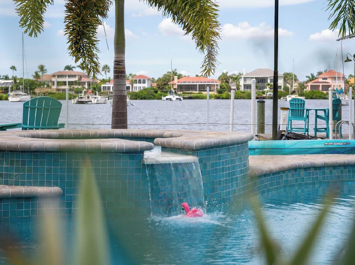 A swimming pool with a waterfall feature, surrounded by palm trees and a view of the marina with boats.