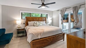 Ein Schlafzimmer mit einem hölzernen Bett, einem Deckenventilator, einem Nachttisch mit Lampe, einem blauen Stuhl und Blick ins Freie durch eine Schiebetür.