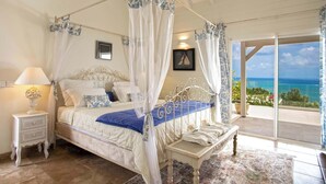 Ein Schlafzimmer mit Himmelbett, Nachttisch, Sitzbank und Meerblick durch eine Glastür.