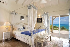 Ein Schlafzimmer mit Himmelbett, Nachttisch, Sitzbank und Meerblick durch eine Glastür.