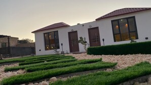A modern two-story building with a white facade and red-tiled roof. There are two large windows on the front, a door, and a small cactus plant in the garden area. - Sun Horizon Chalet (Nizwa)