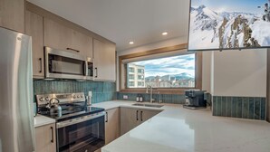 Una cucina moderna con elettrodomestici in acciaio inossidabile, un'ampia finestra che offre vista su edifici e montagne, e un televisore a schermo piatto montato a parete.