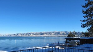 Um deck coberto de neve com guarda-corpo, vista para um lago, uma cabana e montanhas nevadas ao fundo.