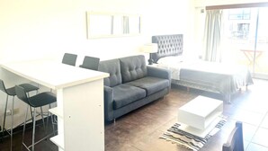 A modern hotel room with a gray sofa, a white desk with two chairs, a bed with a dark headboard, and a large window with curtains.
