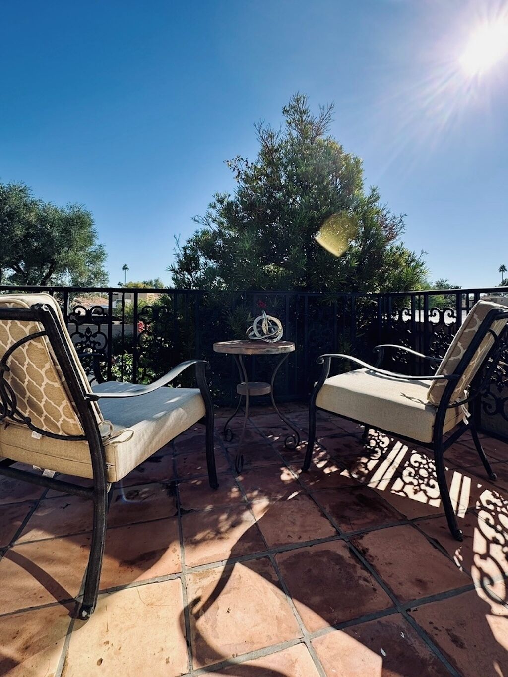 An outdoor patio with two cushioned chairs and a small table, surrounded by a black metal fence and a patterned floor.