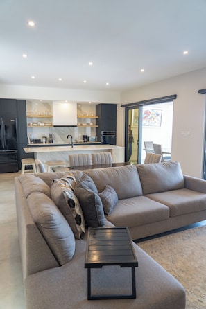 A modern living room with a large sectional sofa, a coffee table, and a kitchen area in the background. - Luxurious Sunset Lagoon View 2-bed bedroom Apartment (Cole Bay)