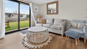 Een woonkamer met een grijze bank, een ronde salontafel en een blauwe fauteuil. Er is een groot raam met uitzicht op een achtertuin en een huis.