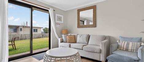 Una sala de estar con un sofá gris, una mesa de centro redonda y un sillón azul. Hay un ventanal con vistas a un patio trasero y una casa.