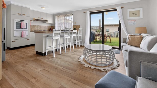 Een moderne keuken met witte kasten, een eethoek met witte stoelen, een ronde salontafel en uitzicht op een achtertuin door een schuifdeur van glas.