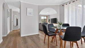 Een moderne woonkamer met een eethoek, met een glazen tafel, donkere stoelen en een flatscreen televisie.