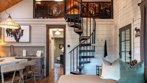 A rustic cabin interior with a wooden staircase, a dining area with bar stools, and a bed with pillows. - Upscale Designer Log Cabin, Cozy, Hot Tub, Firepit (Driggs)