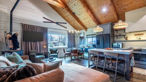 A spacious living area with a high ceiling, wooden beams, a large sofa, a dining area with bar stools, and a kitchen with modern appliances. - Upscale Designer Log Cabin, Cozy, Hot Tub, Firepit (Driggs)