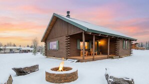 A log cabin with a fire pit in the foreground, a covered porch, and a snow-covered landscape. - Upscale Designer Log Cabin, Cozy, Hot Tub, Firepit (Driggs)