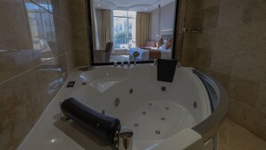 A modern bathroom with a large bathtub, a mirror, and a view of a bedroom with a bed and curtains. - Armada Residence Hotel (Taif)