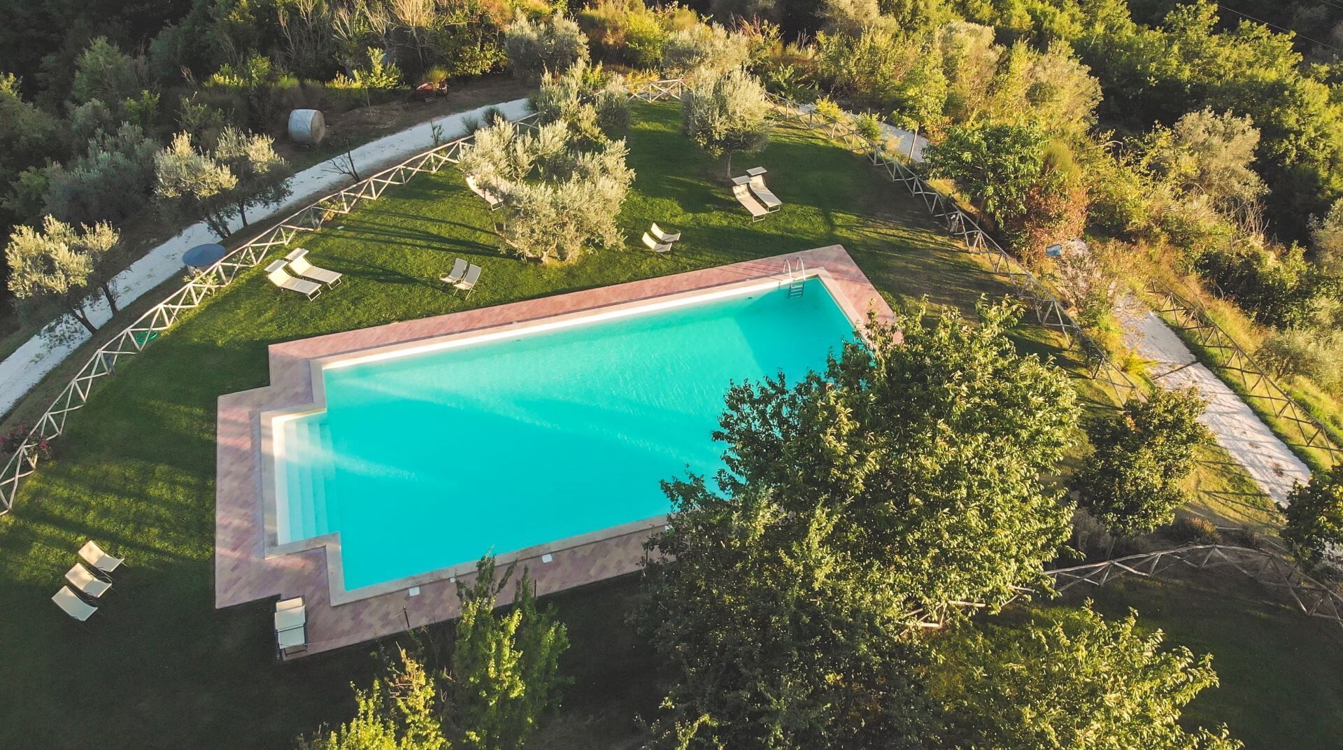 An aerial view of a rectangular swimming pool surrounded by a wooden deck, with lounge chairs and a walking path.