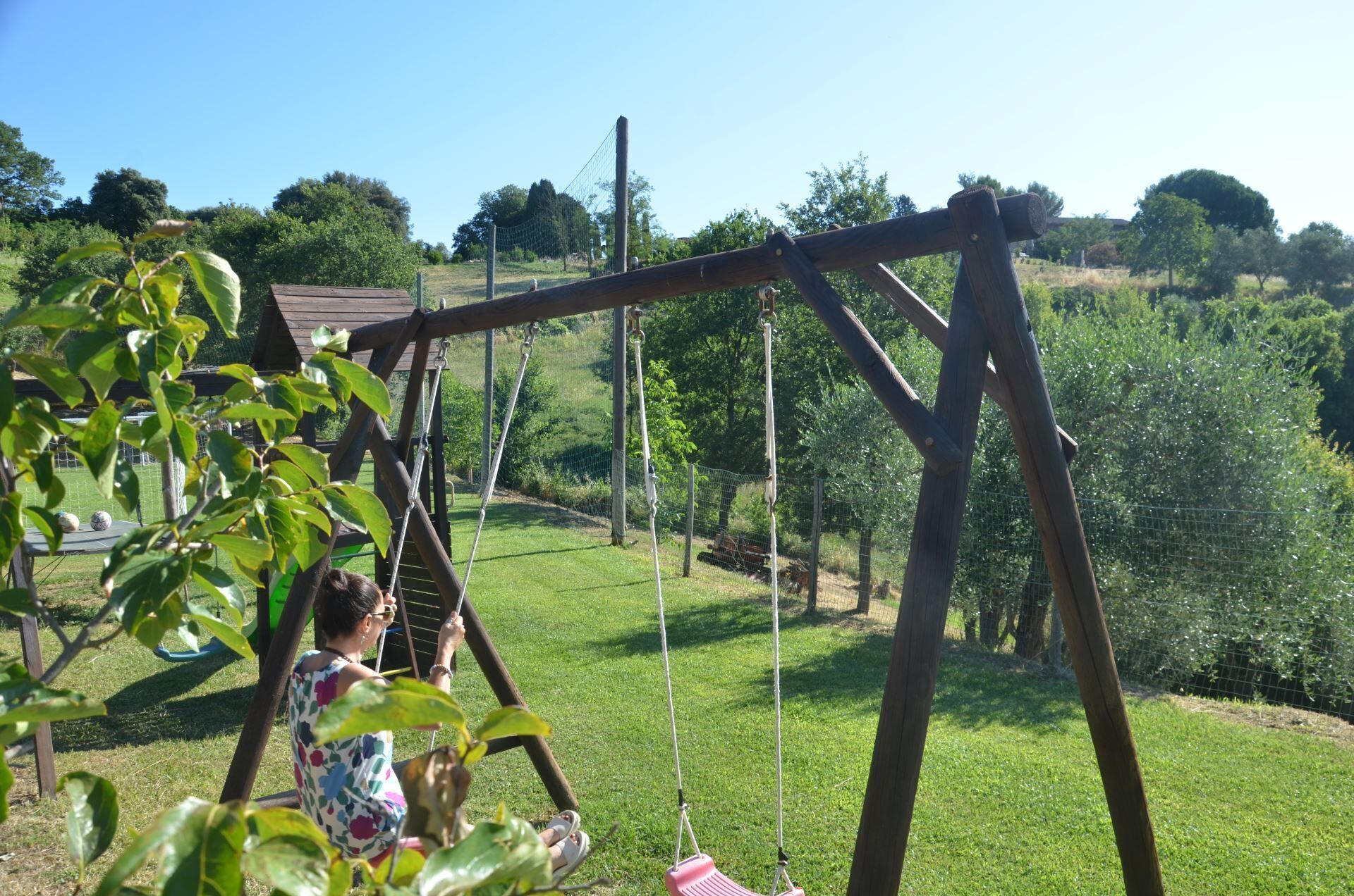 Un ensemble de balançoires en bois installé dans un espace herbeux, offrant une vue dégagée sur les arbres et une clôture en arrière-plan.