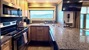 Eine moderne Küche mit Granit Arbeitsplatten, Edelstahlgeräten, Holzschränken, einem großen Fenster mit Blick und einem flachbildschirmfernseher über der Spüle.