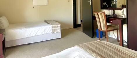A hotel room with a bed, a desk, a chair, a ceiling fan, a painting, and a door leading to another room.