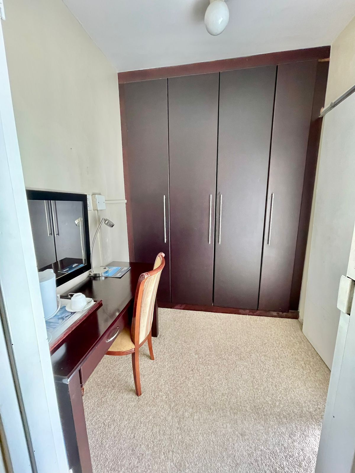 A room with a dark wooden wardrobe, a desk with a computer and lamp, and a chair.