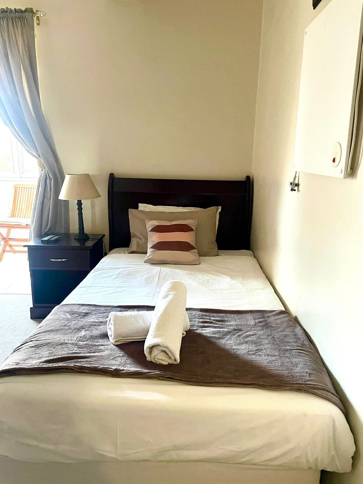 A neatly made bed with a rolled towel, a bedside lamp, and a nightstand with a remote control.