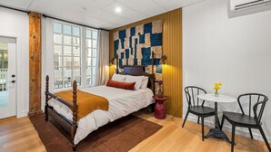 Une chambre avec un lit en bois, une grande fenêtre, un climatiseur fixé au mur et une petite table ronde avec des chaises.