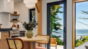 Una cocina moderna con comedor, armarios de madera, un amplio ventanal con vista al océano y una lámpara colgante.