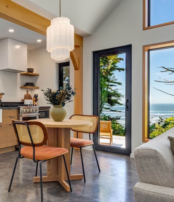 A modern kitchen with a dining area, wooden cabinets, a large window with a view of the ocean, and a hanging light fixture. - Fogged Inn Cabin (Ucluelet)