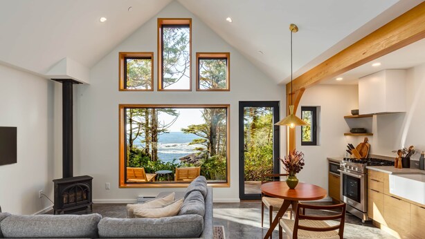 A modern living room with a large window overlooking the ocean, a cozy seating area, and a kitchen with wooden beams. - Fogged Inn Cove (Ucluelet)