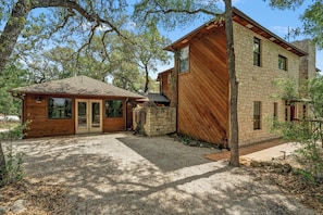 A two-story wooden house with a stone wall, a gravel driveway, and surrounding trees.