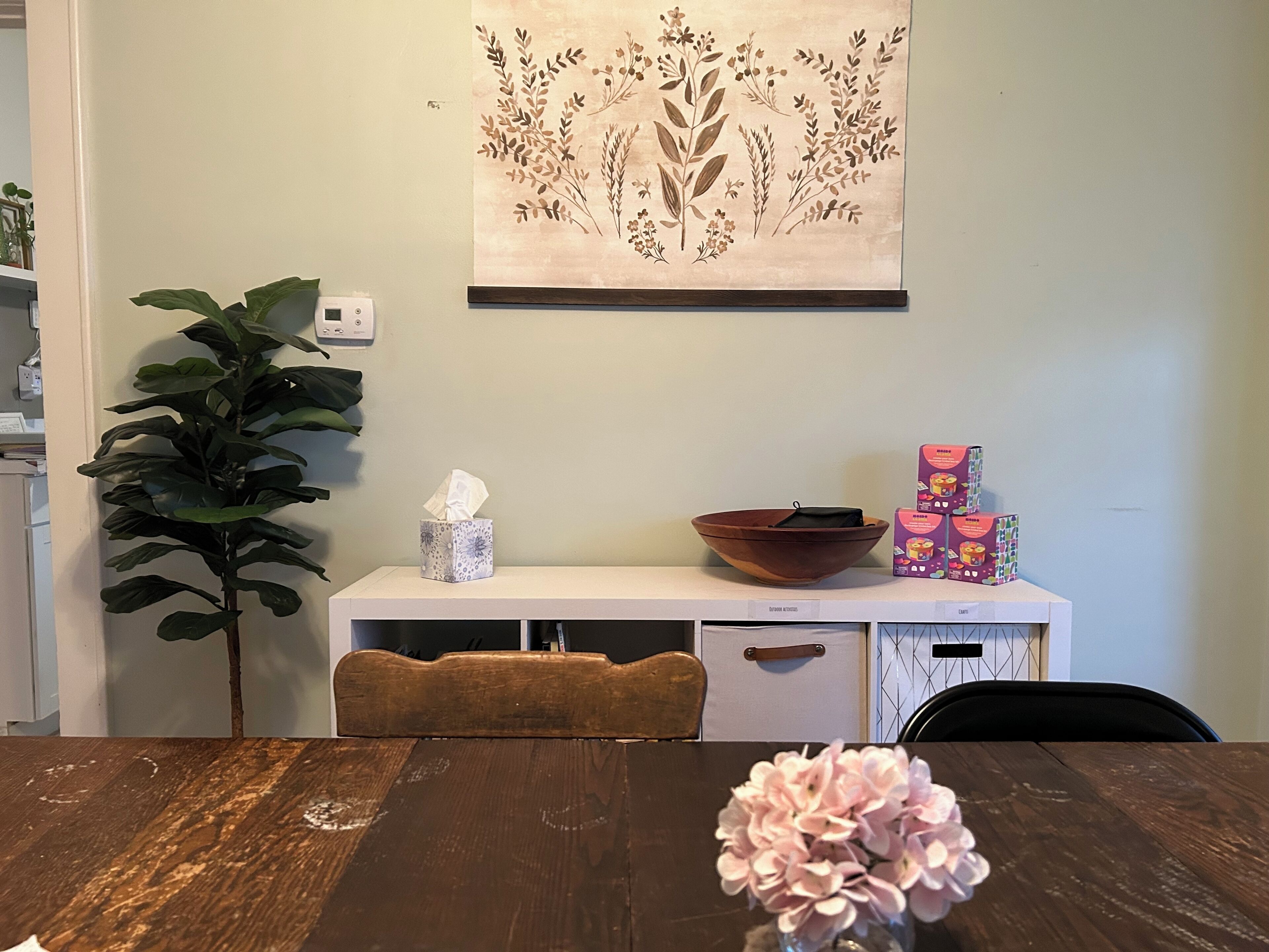 A dining area with a wooden table, a vase of flowers, a plant, a wall-mounted artwork, a white cabinet, and a box of tissues.