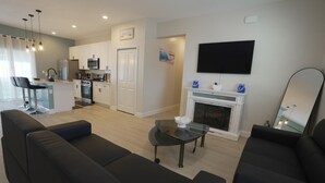 Een moderne woonkamer met een zwarte bank, een glazen salontafel en een open haard. Er is een keuken met witte kasten en een eethoek met een bar.