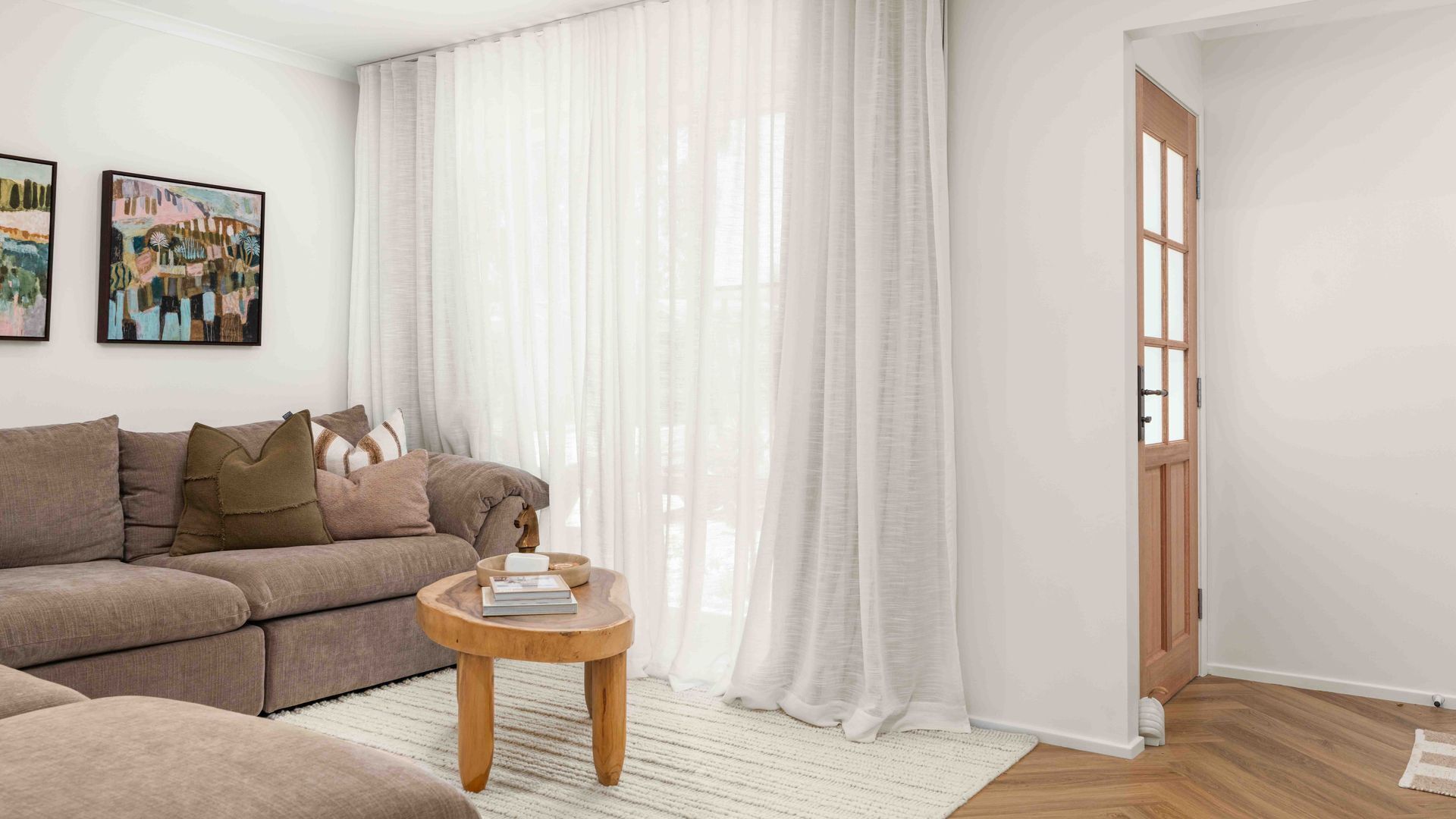 A modern living room with a beige sofa, a round wooden coffee table, and a large window with sheer curtains.