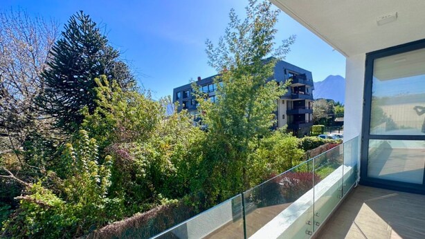 A balcony with a glass railing overlooking a garden and modern buildings. - Pucón CENTRO - Steps away from Casino ENJOY - Apartment NEW - A/C - Volcano View (Pucon)