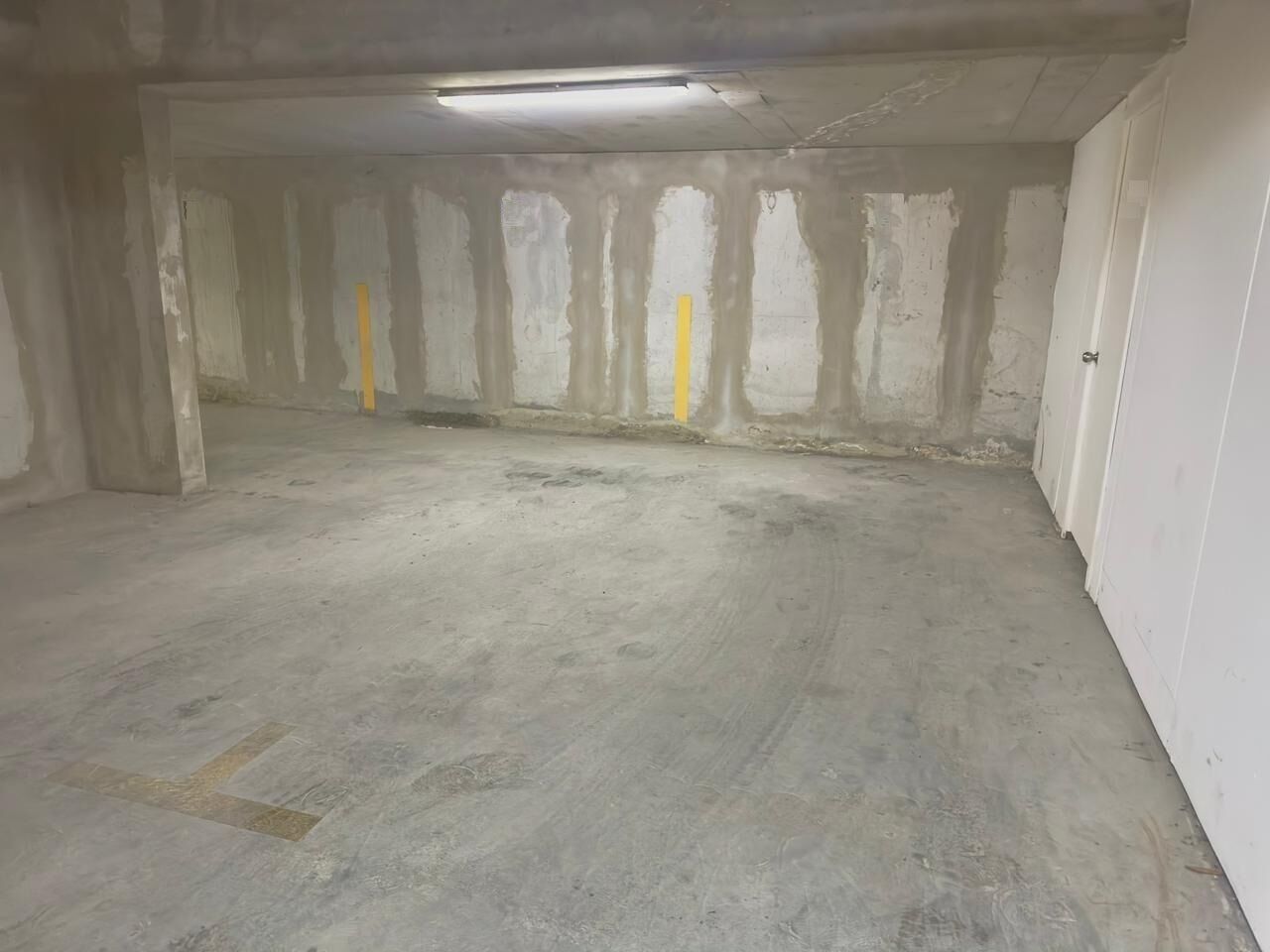 Un espacio amplio e incompleto con paredes y suelo de hormigón, una lámpara fluorescente y una puerta a la derecha.