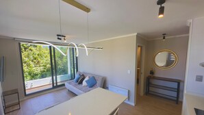 A modern kitchen with a large island, a dining area with a table and chairs, a sofa with cushions, and a view of greenery through the windows. - Pucón CENTRO - Steps away from Casino ENJOY - Apartment NEW - A/C - Volcano View (Pucon)