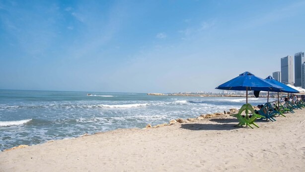 Ein Strand mit blauen Sonnenschirmen und Liegestühlen, ein sandiger Uferstreifen und Blick auf den Ozean.