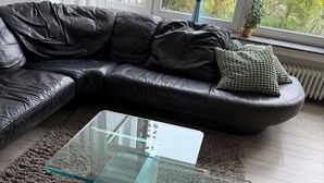 A modern living room with a glass coffee table, a black leather sofa, and a view of greenery through the window.