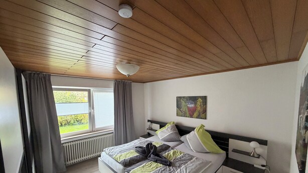 A bedroom with a wooden ceiling, a bed with grey and green bedding, a window with curtains, a radiator, and a painting on the wall.