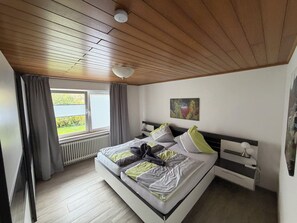 Une chambre avec un plafond en bois, un lit avec une literie grise et verte, une fenêtre avec des rideaux, un radiateur et un tableau accroché au mur.