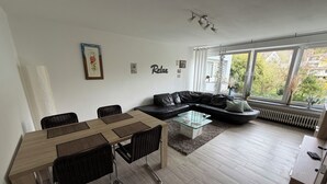 A modern living room with a dining area, a black leather sofa, a glass coffee table, and a large window with a view of greenery.