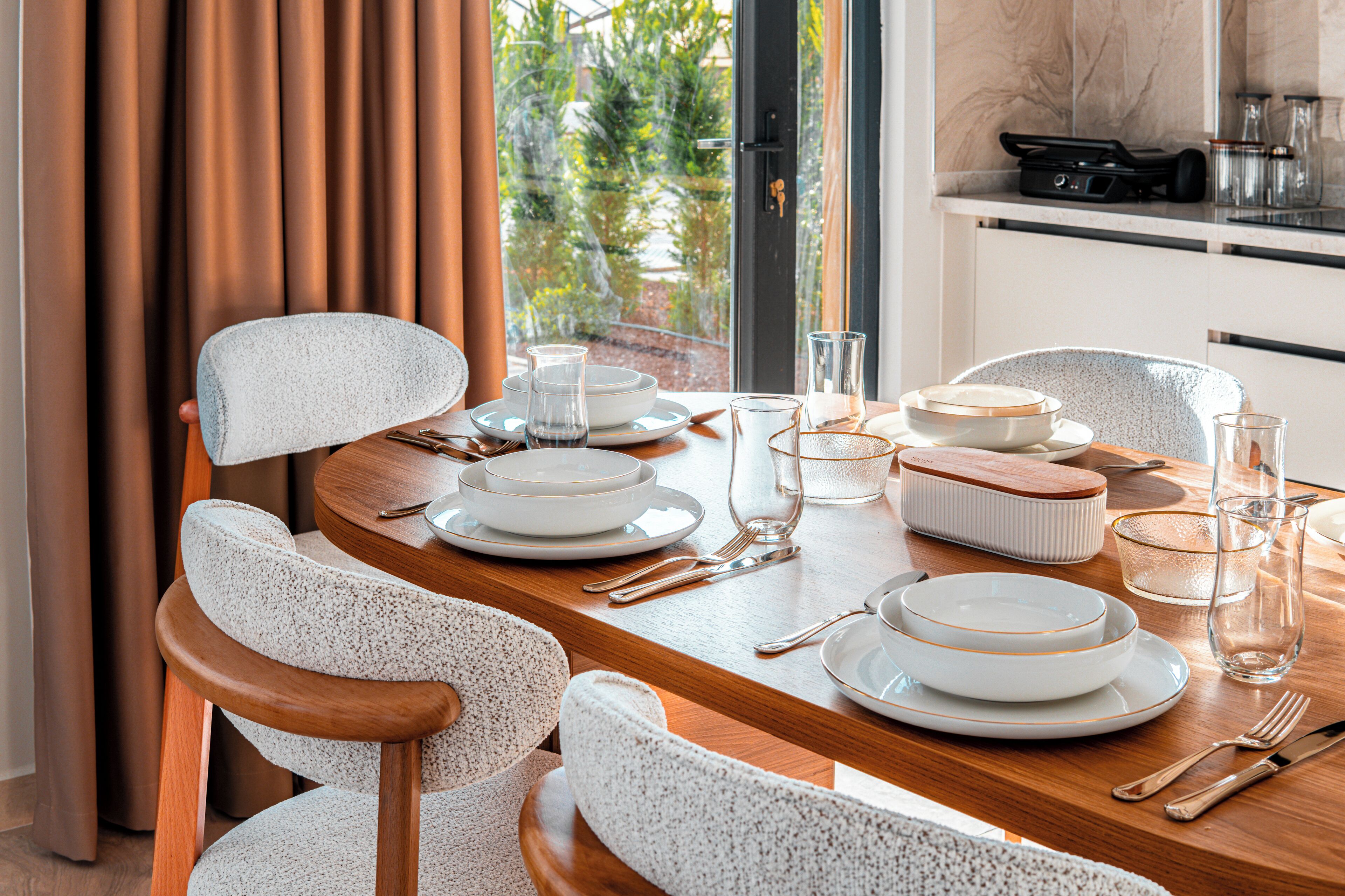 A dining table set with plates, glasses, and cutlery, with a view of greenery outside.