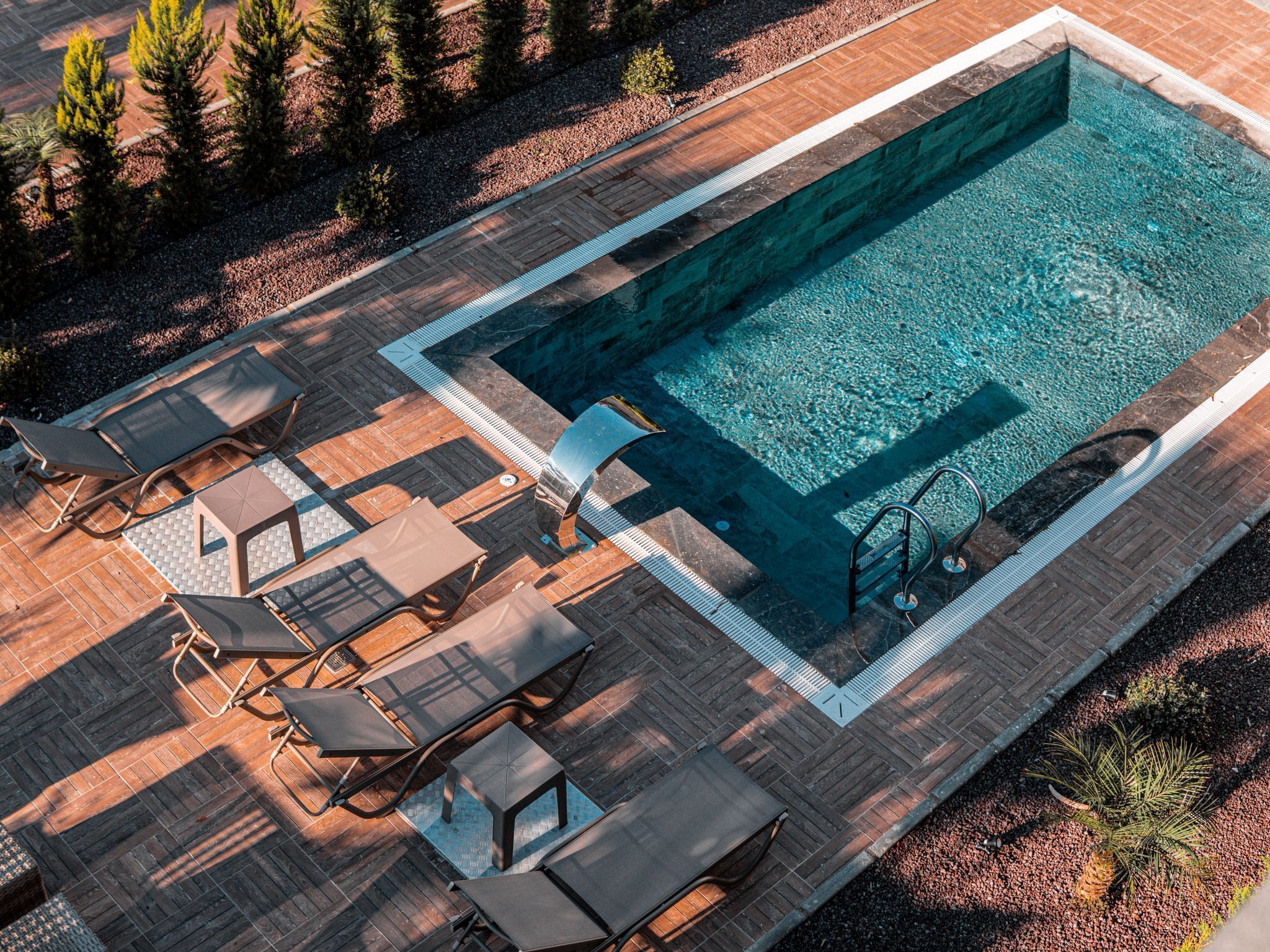 Ein Luftbild eines rechteckigen Schwimmbeckens mit klarem, blauem Wasser, umgeben von einer Holzterrasse mit Liegestühlen und einem kleinen Tisch.
