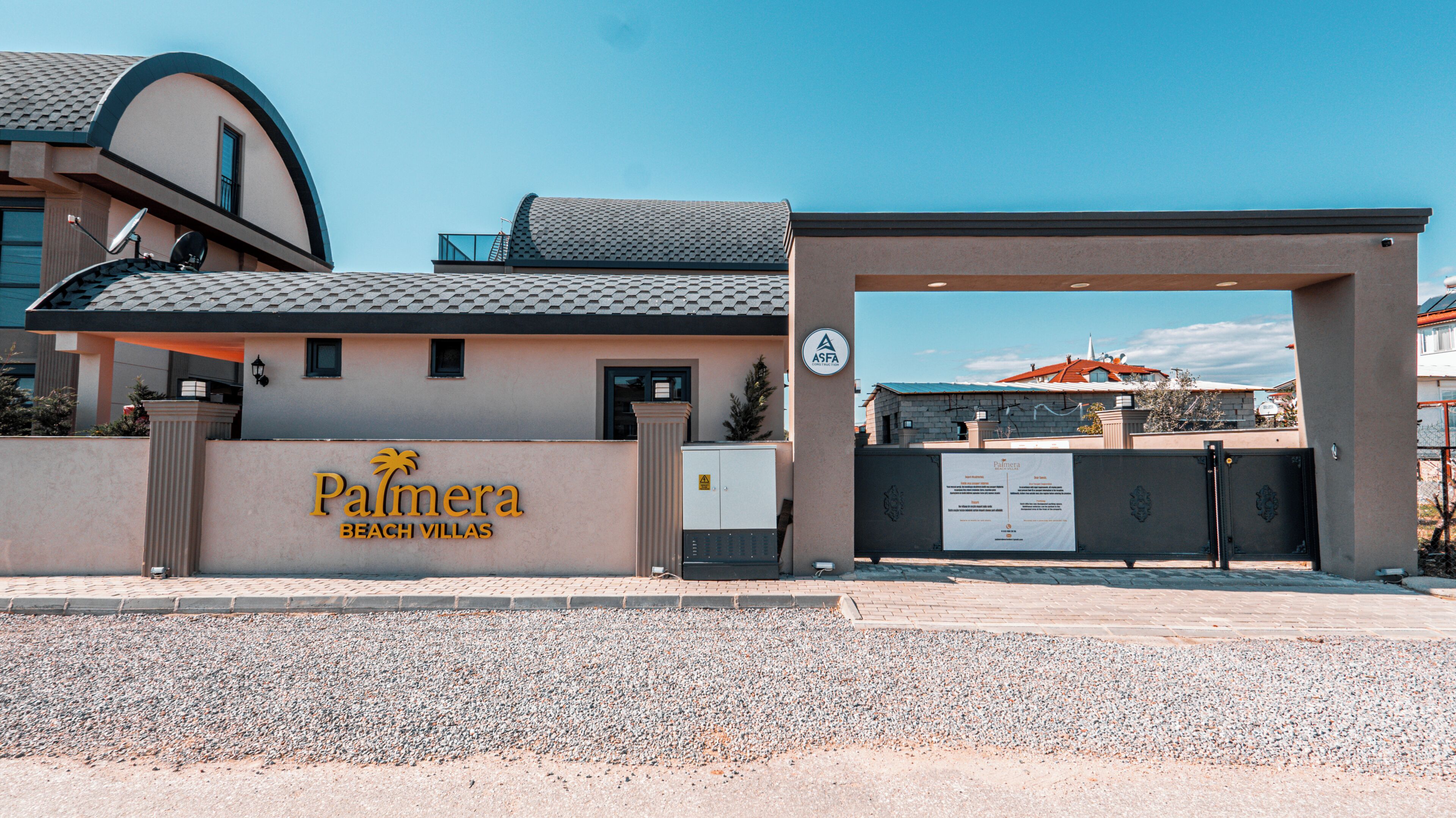 Una entrada cerrada a una propiedad llamada 'Palmera Beach Villas', con cielo despejado y un edificio con tejado de tejas al fondo.