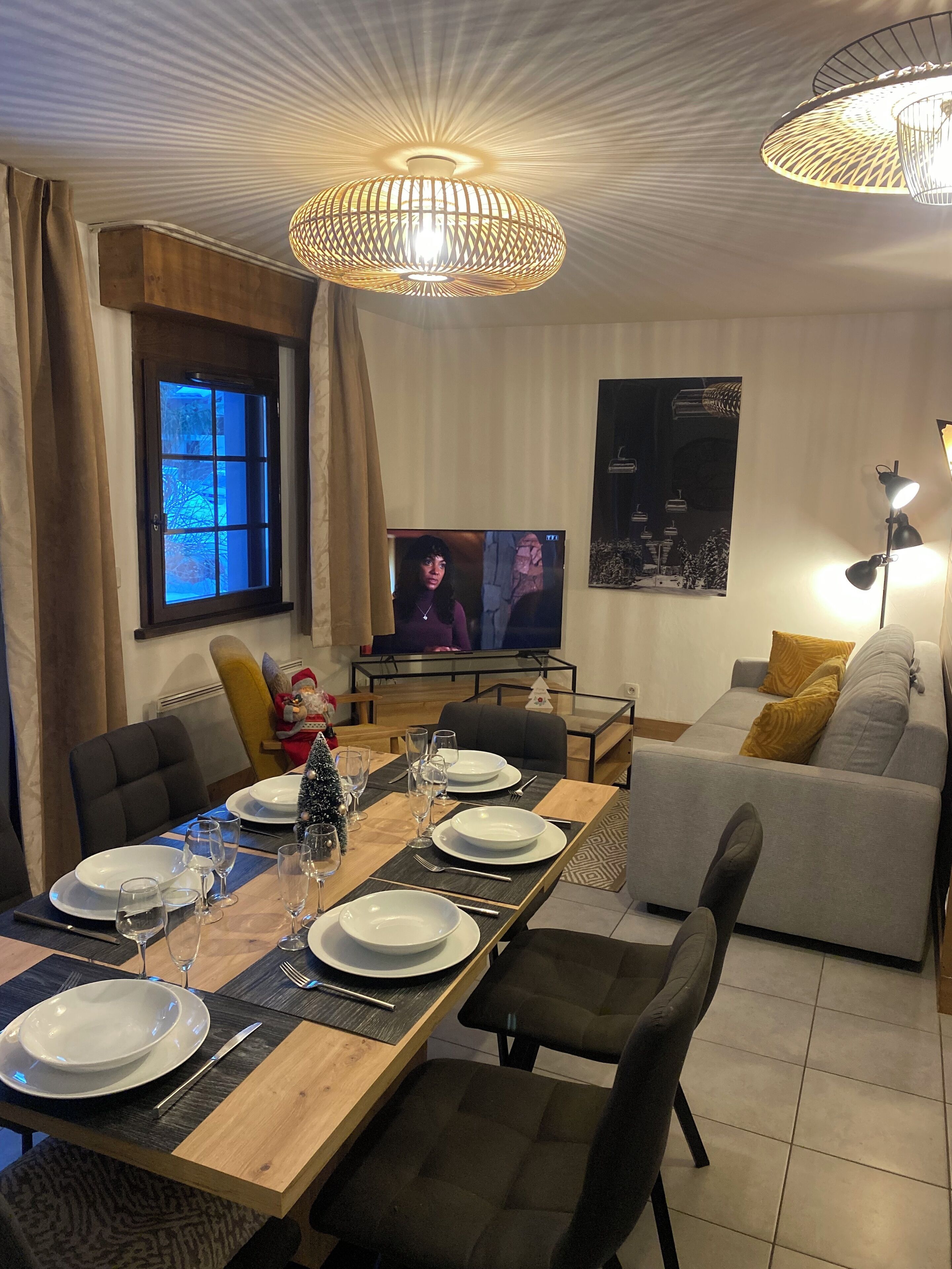 Een eetruimte met een lange houten tafel voor zes personen, een grijze bank en een televisie op de achtergrond.