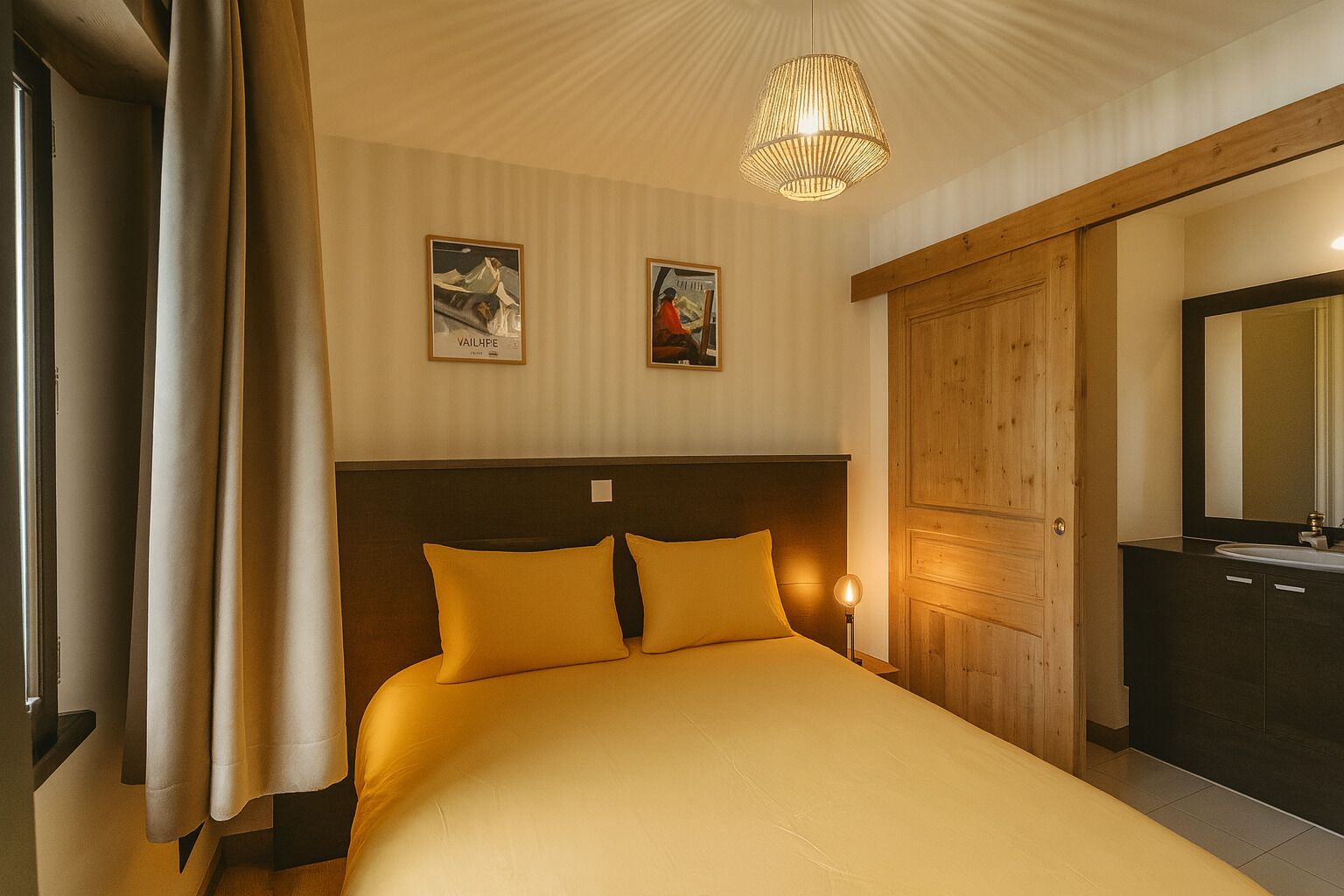 A hotel room with a bed, a wooden wardrobe, a mirror, and two framed pictures on the wall.