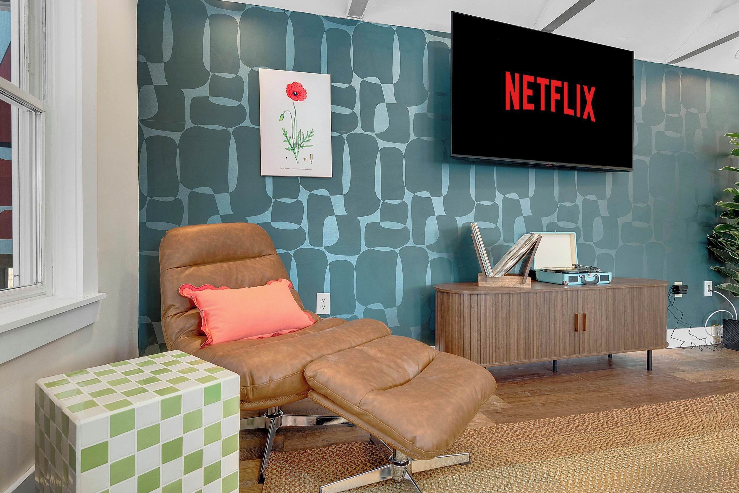 Una sala con un sillón de cuero marrón, un reposapiés blanco y verde a cuadros, un aparador de madera y un televisor montado en la pared mostrando 