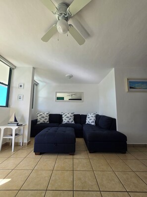 A living room with a sectional sofa, a ceiling fan, and a small white table with a model ship. - Beach-5 Pools-25 Min from airport-Unit with reserve water system  (Loíza)